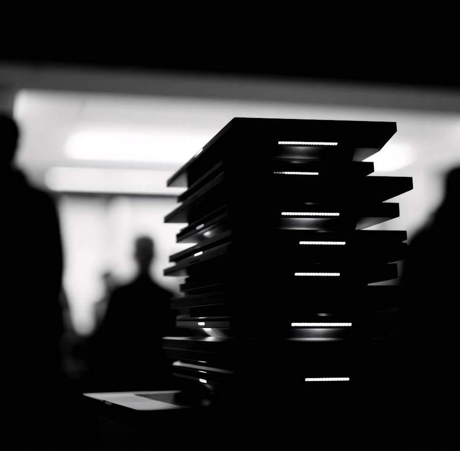 Black-and-white image of a stack of illuminated objects with blurred silhouettes in the background, creating a modern and abstract vibe.